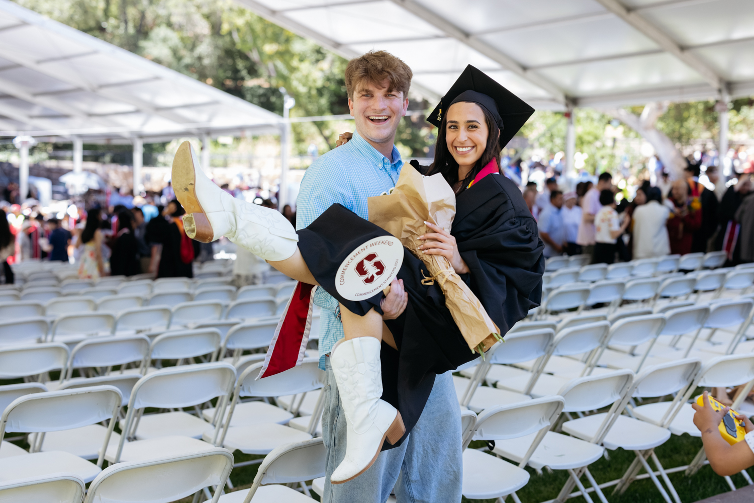 A graduate in a cap and gown is being playfully carried by a friend, both smiling at a graduation ceremony.
