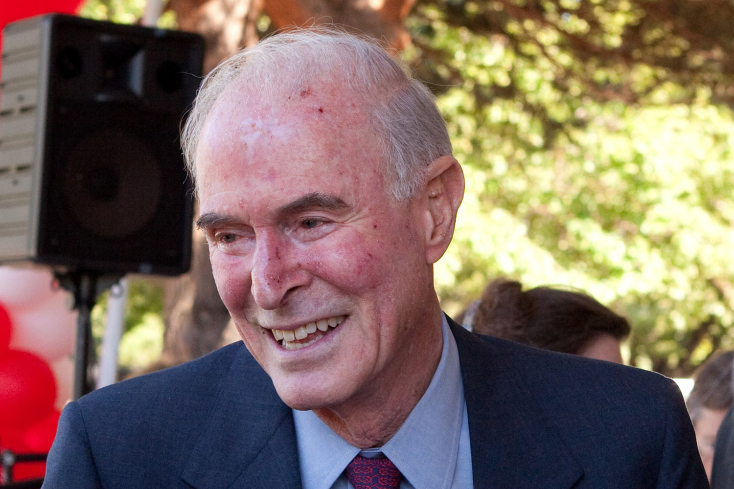 Smiling, candid shot of Peter Bing in a navy suit outdoors with trees in the background.