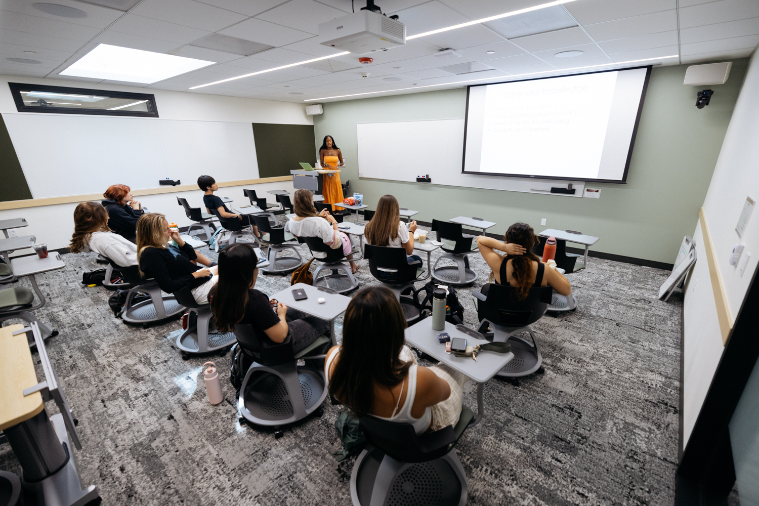 Gabby Edelin stands in front of a classroom filled with students seated at desks, engaging them in a lesson projected on a screen.