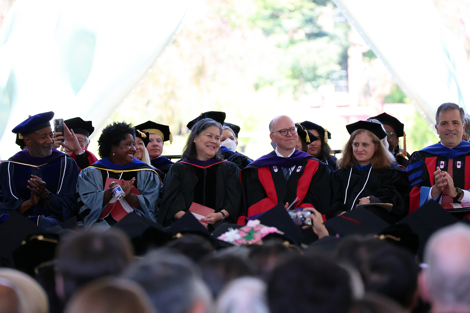 Faculty members in academic robes sit together, applauding during a commencement ceremony with a crowd of graduates in view.