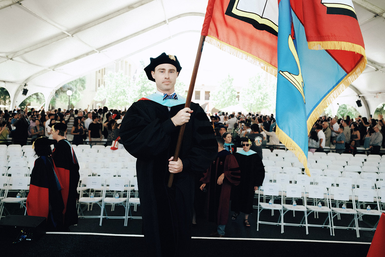 PhD grad Brendan Fereday in academic dress proudly holds a large flag while standing in front of a crowd during a graduation ceremony.