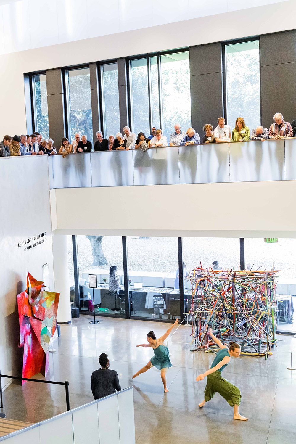 Dancers perform in art gallery while an audience watches from a balcony above, with colorful art installations nearby.