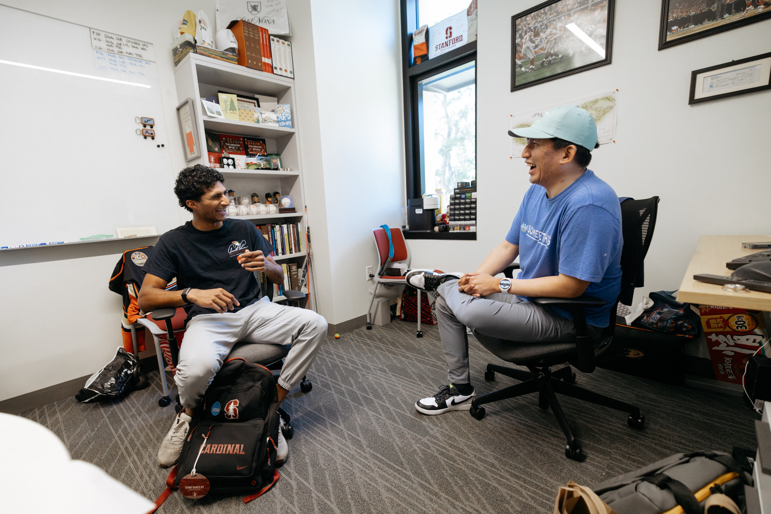 Samir Banerjee laughing during a conversation with one of his teammates in a Stanford office with Cardinal athletics photos and paraphernalia in the background.