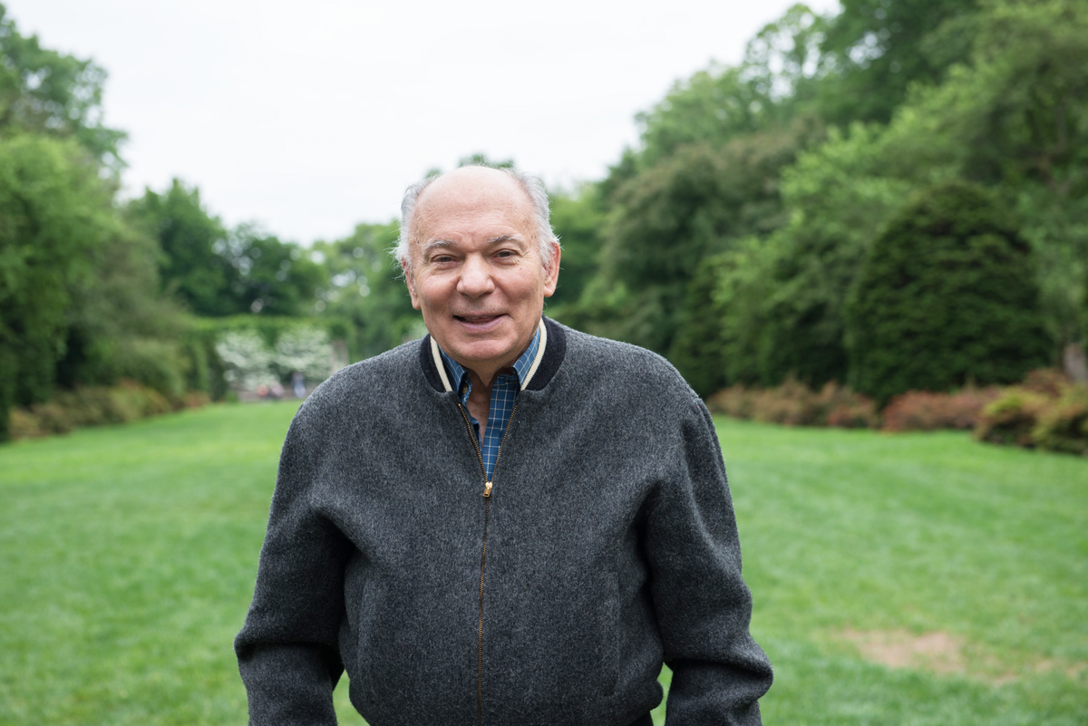 Rosaldo in a gray sweater is smiling and standing in a garden, surrounded by trees and plants.
