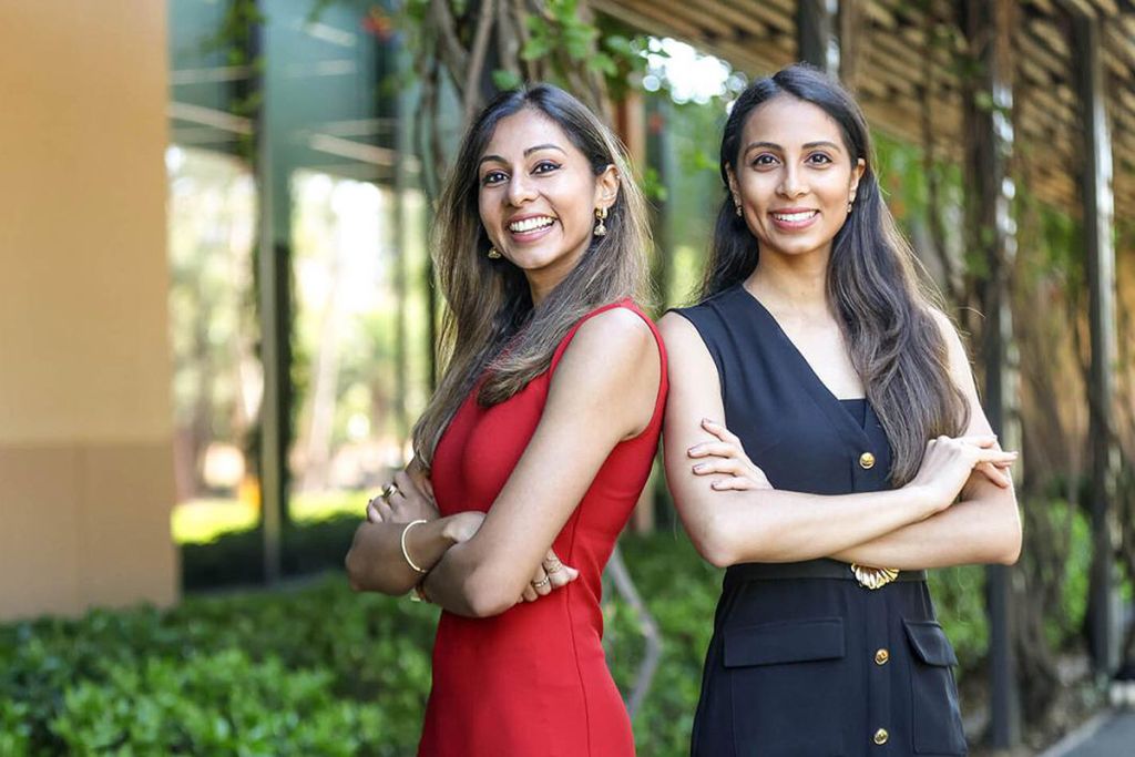 Two women stand confidently outdoors, smiling and posing with their arms crossed, wearing dresses in red and navy.