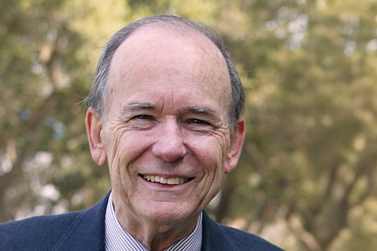 A smiling older man in a suit stands outside, surrounded by greenery.