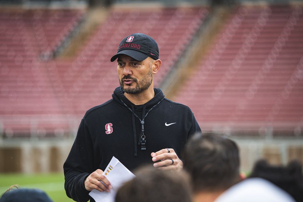Stanford coach in a black cap and hoodie, holding papers in one hand, speaks to a group of players in an empty stadium.