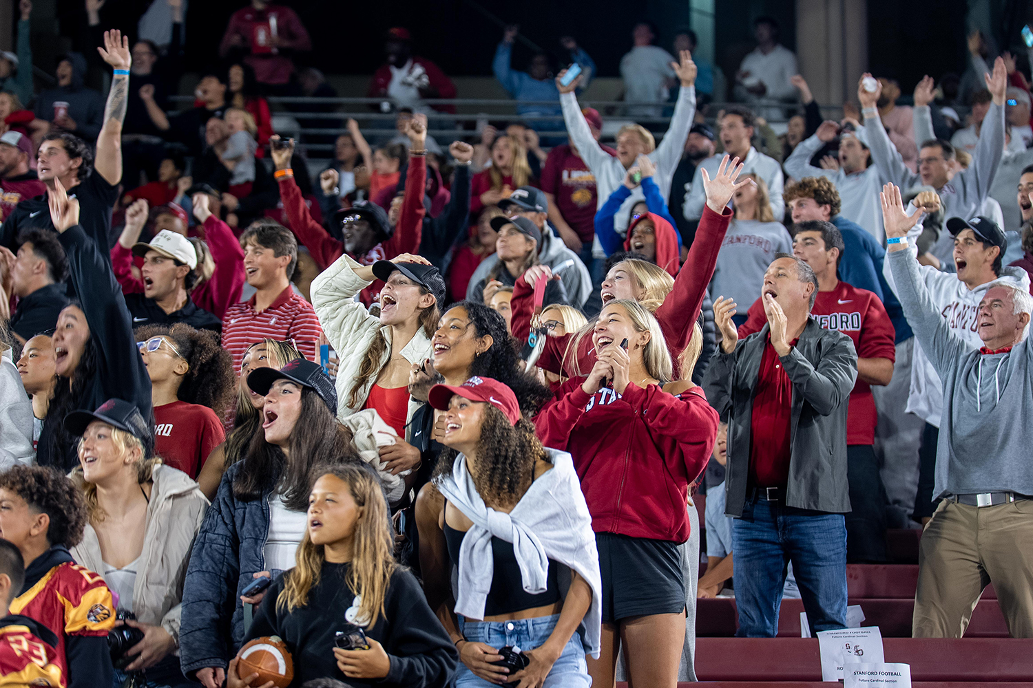 A crowd of Stanford fans cheers with raised arms during a night game.