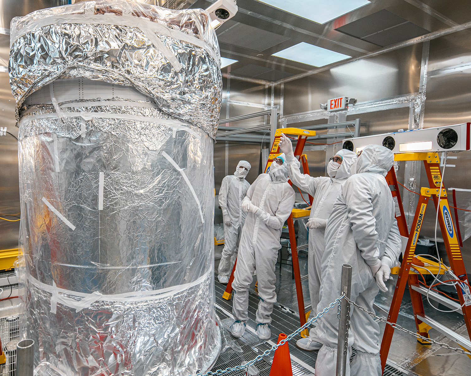 Researchers in white clean suits gather around a tall detector wrapped in foil and plastic inside a clean room.