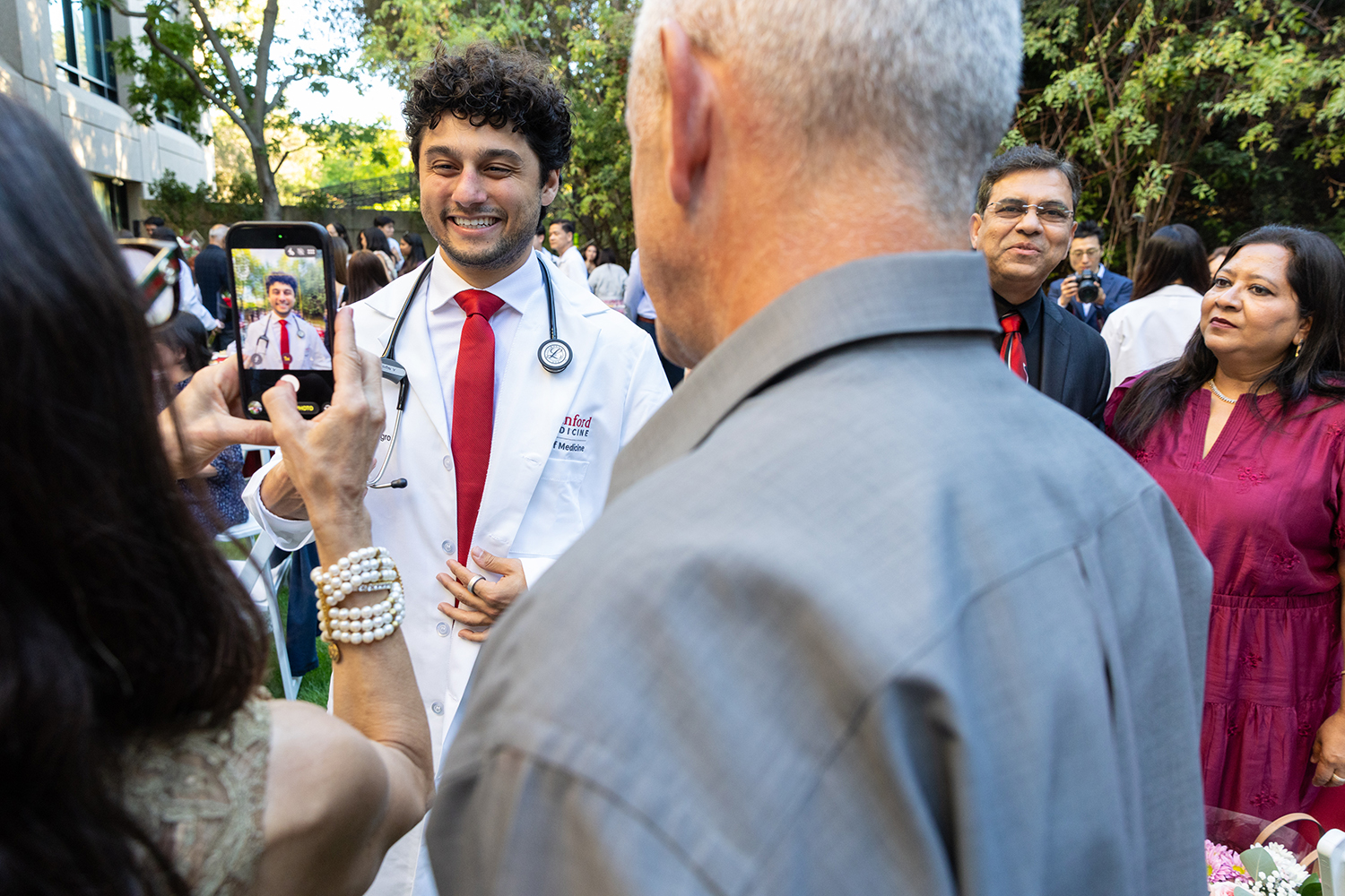 A medical student in a white coat smiles as a family member takes his photo at an outdoor celebration.