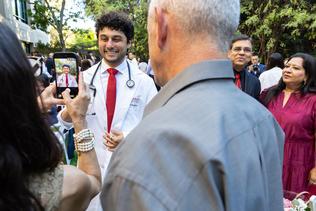 A medical student in a white coat smiles as a family member takes his photo at an outdoor celebration.