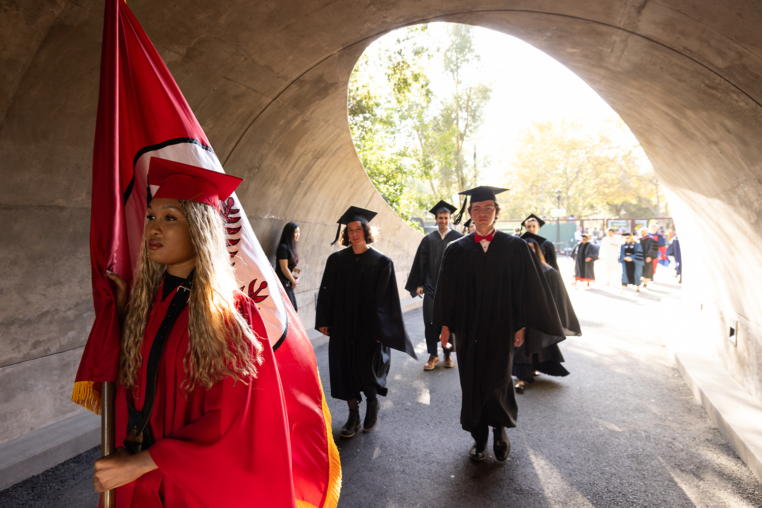 A flag bearer in a red academic gown leads a procession through a stone tunnel toward the ceremony.