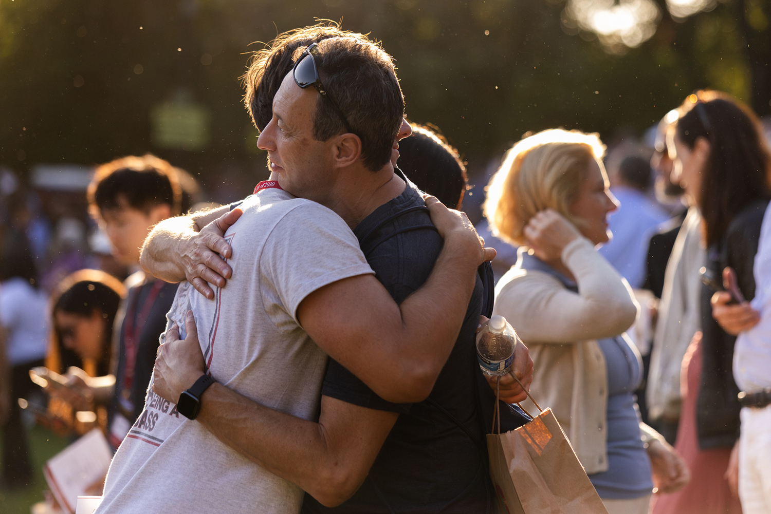 A father embraces his student warmly at an outdoor gathering, surrounded by other attendees.