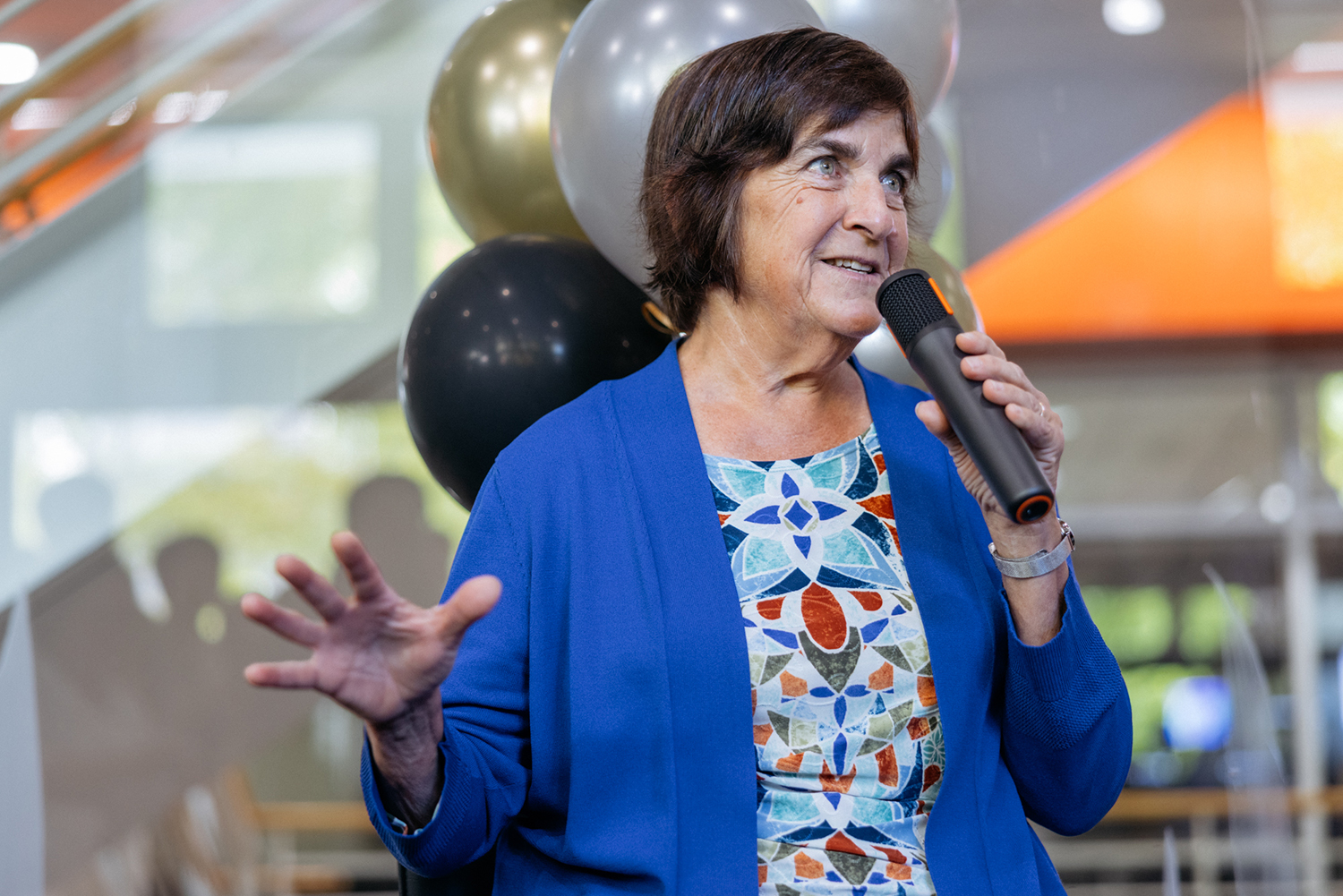 Jennifer Widom speaks into a microphone in front of black, gold, and silver balloons.