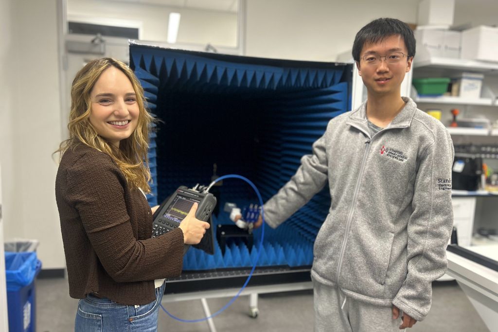 Two researchers pose beside a foam-lined test chamber, one holding a handheld signal analyzer connected by a blue cable.
