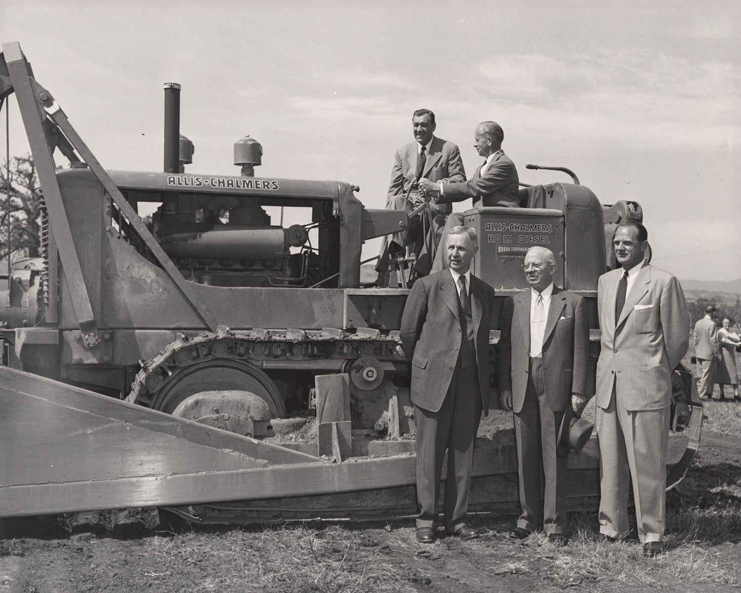  Former Stanford president J. E. Wallace Sterling, top left, and others posing with an Allis-Chalmers bulldozer in an open field.