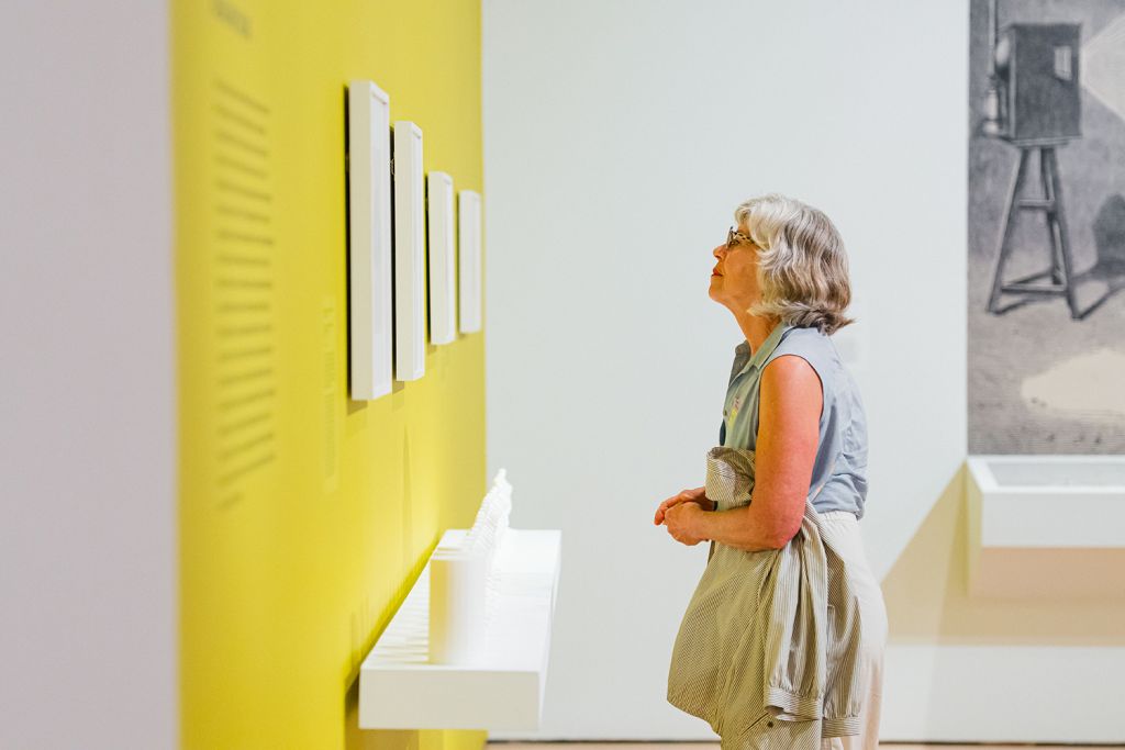 Museum visitor looks up at a row of small framed works on a bright yellow gallery wall.