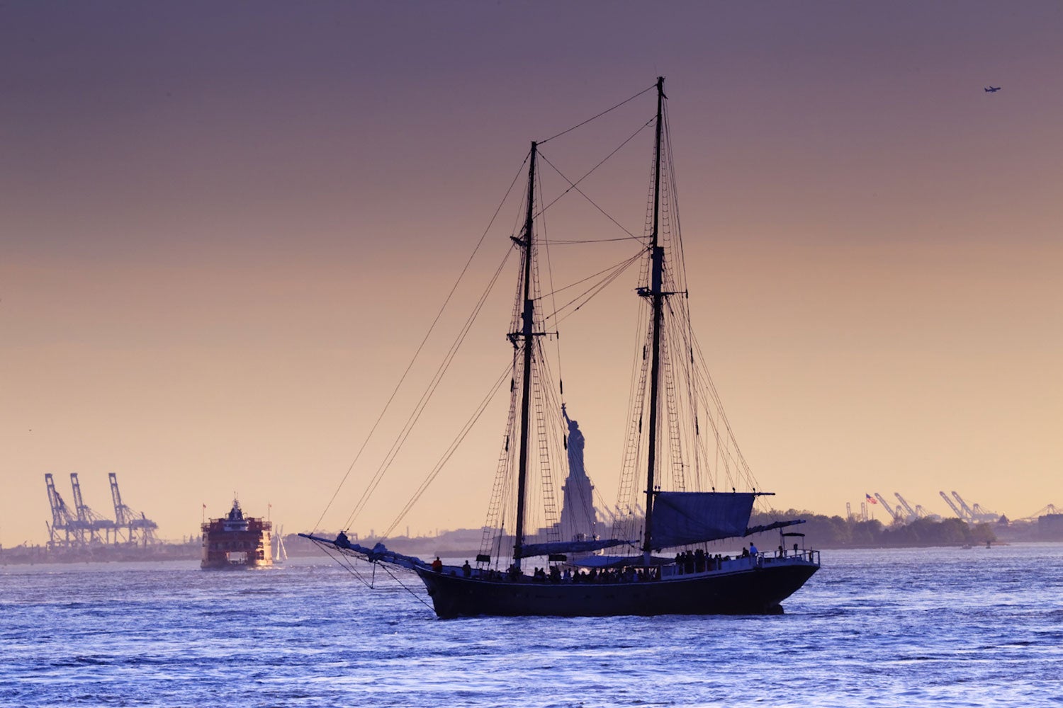 A recreational sailboat in front of the Statue of Liberty, with signs of industry in the background.