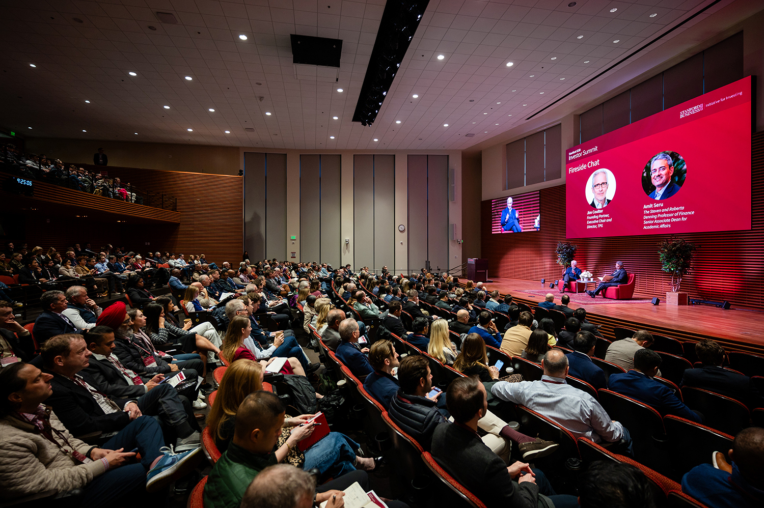 A large audience attends a fireside chat event featuring two speakers on stage, with a large screen displaying their names and topics.