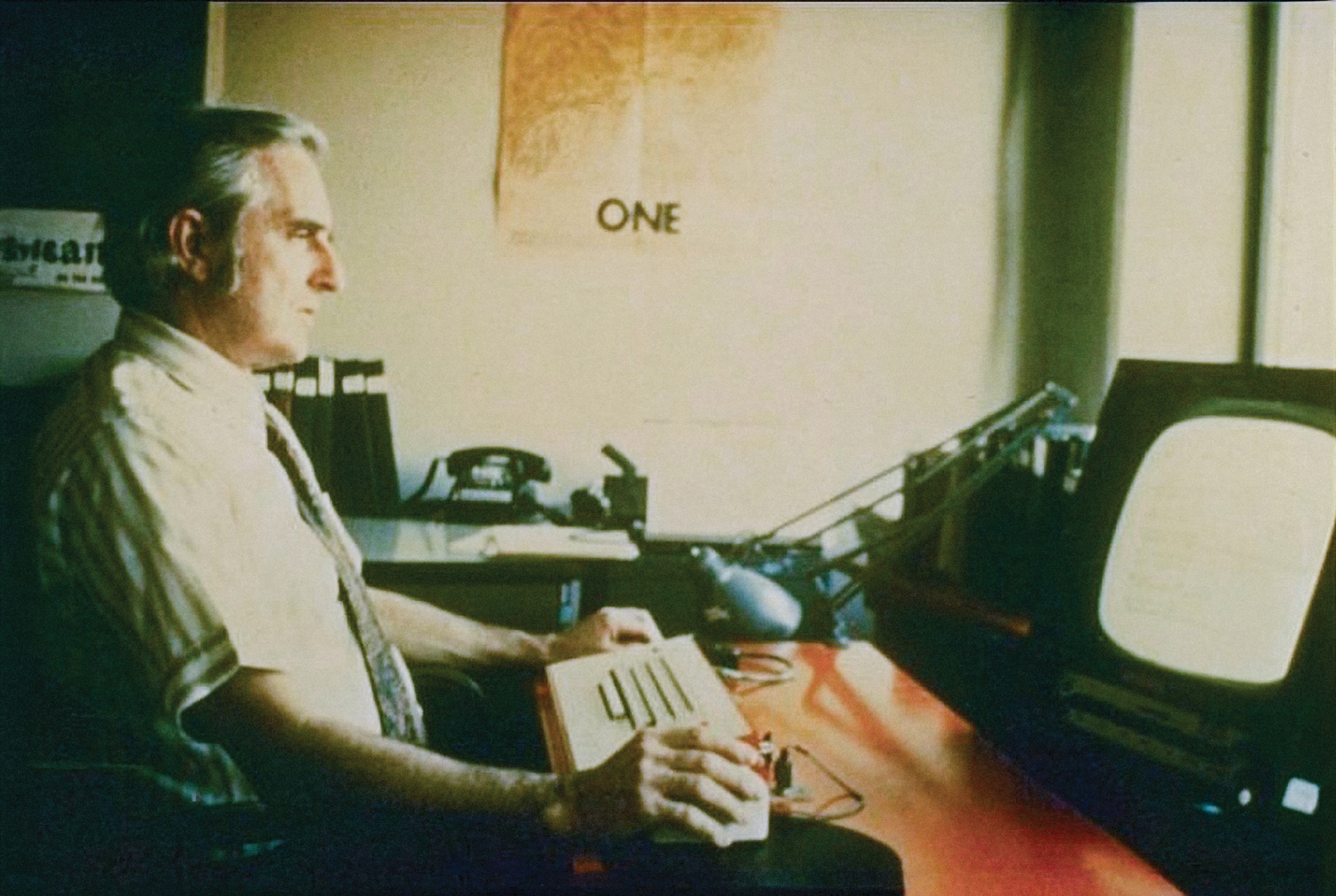 Doug Engelbart in shirt and tie sitting at a desk using a mouse with early computer equipment.