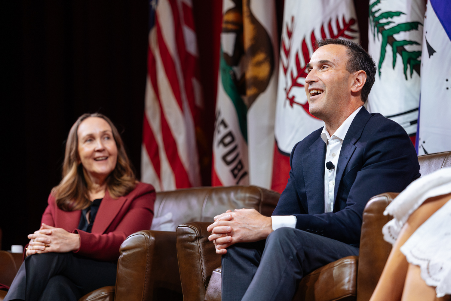 The president and provost seated on couches during a panel discussion, with flags in the background and a focus on their engaged expressions.