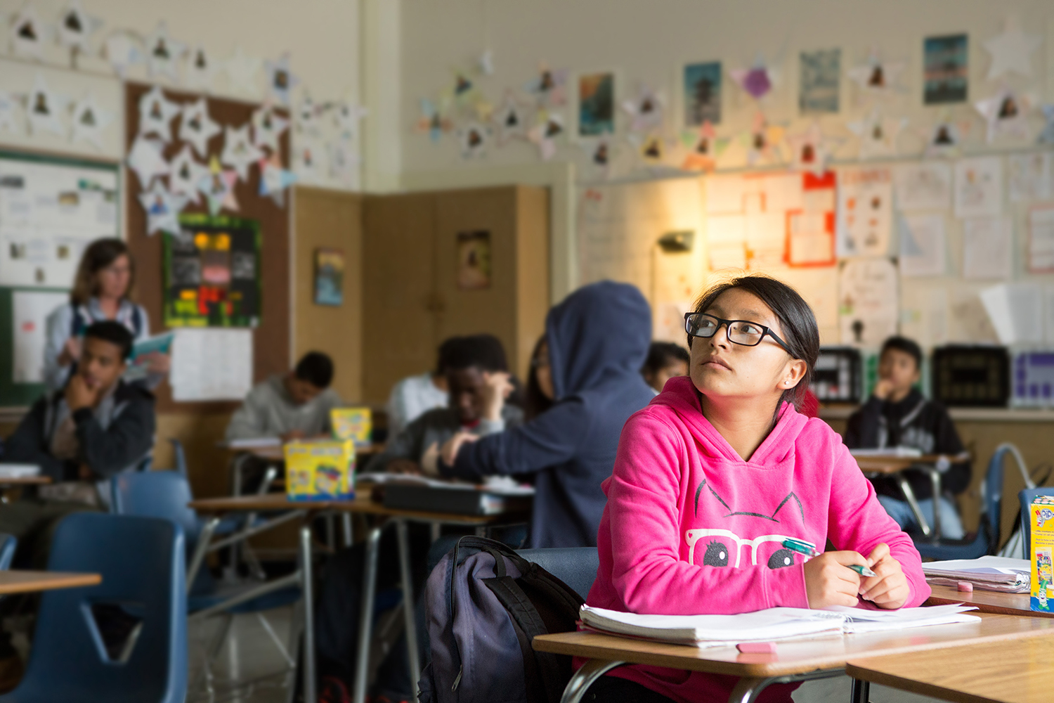 Students are seated in a classroom, with one girl in a pink hoodie actively engaged at her desk.