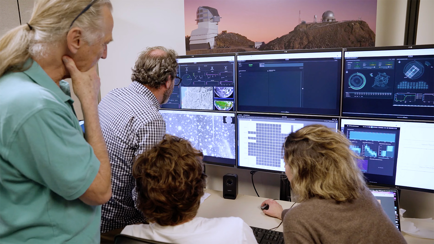 A group of four people studies data on multiple computer screens in an office setting, with an observatory visible in the background.