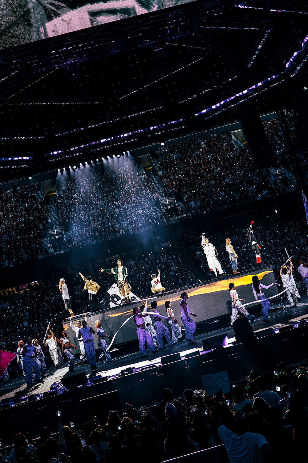 BTS dance and sing on stage during a concert, with a large enthusiastic audience and vibrant lighting in the background.