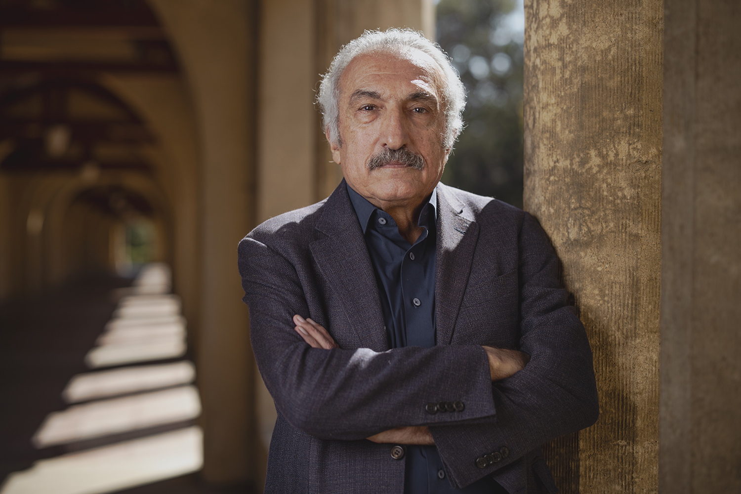 Milani with white hair and mustache in dark blazer standing with arms crossed in a campus arcade.
