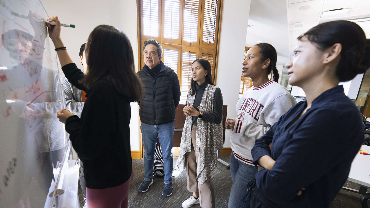 A group collaborates in a meeting room, with one person writing on a whiteboard while others listen and discuss ideas together.