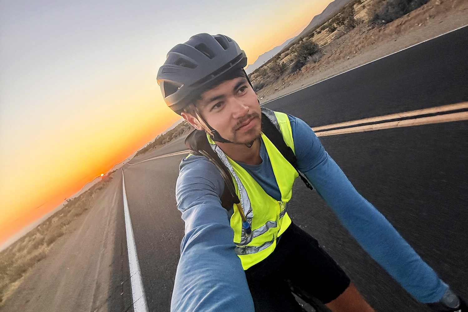 A cyclist wearing a reflective vest rides on a road at sunrise, with colorful skies and mountains in the background.
