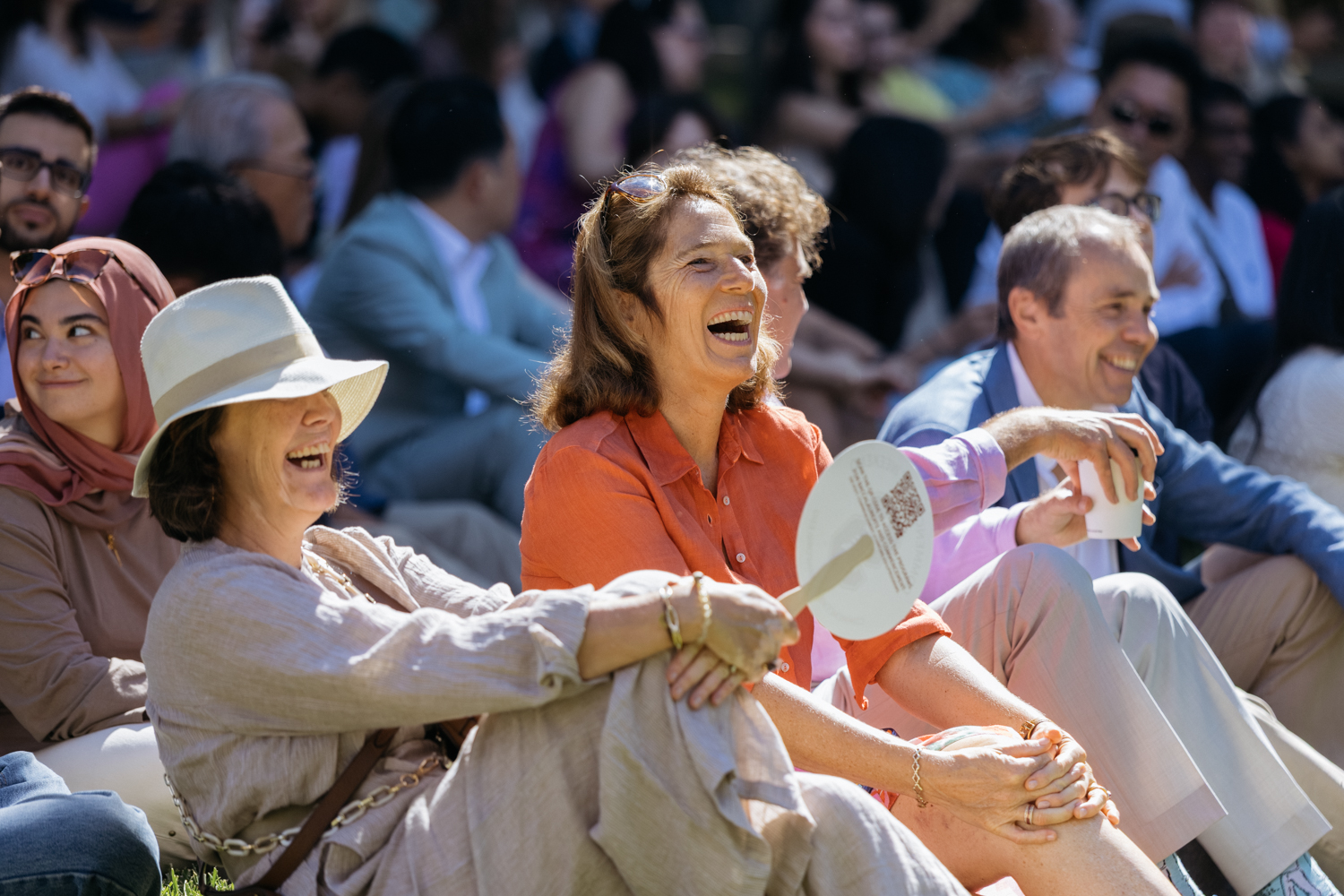 Two women sit outdoors during an event, one holding a fan, as an audience gathers behind them.