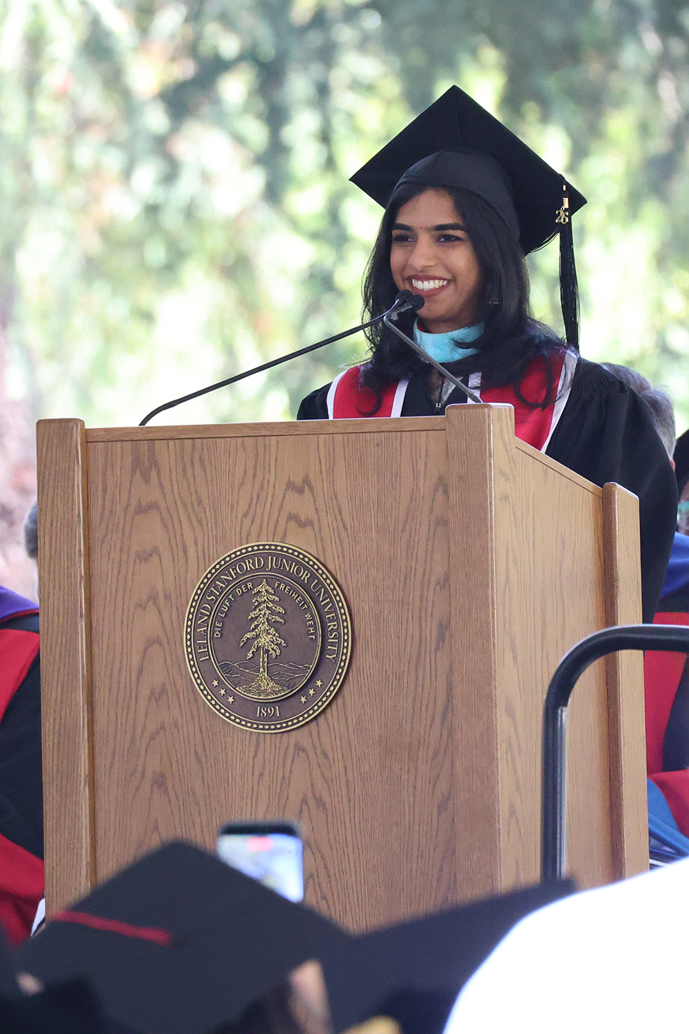 Graduate Mahishaa Balraj in a cap and gown speaks at a podium during a commencement ceremony, with a crowd of fellow graduates visible in front.
