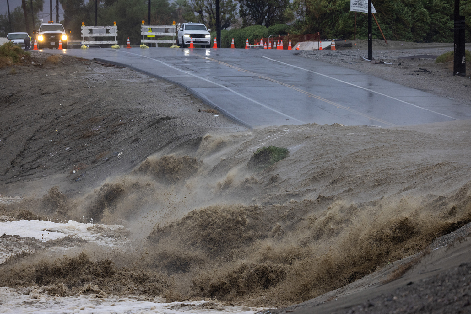 Floodwaters surge over a road and embankment during heavy rainfall.