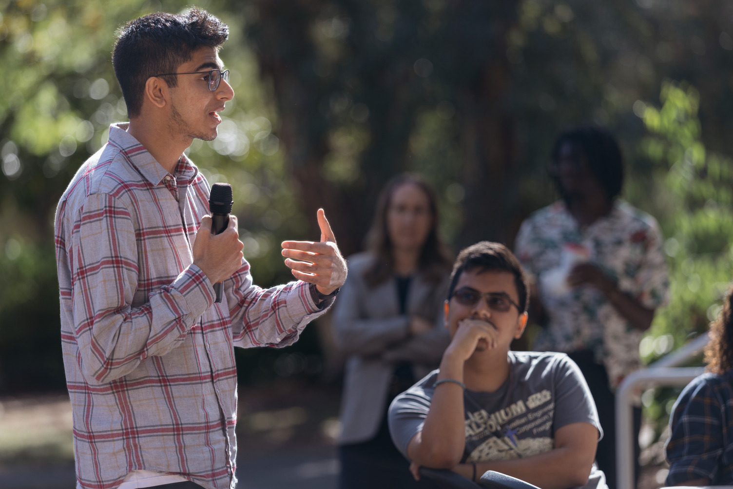 A student with microphone gestures while speaking to an outdoor audience of seated listeners.