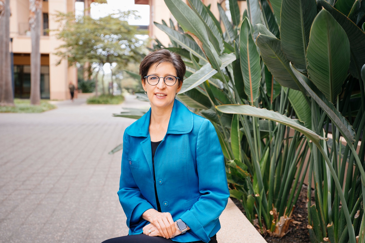 Open video Susan Athey sitting on a bench near tropical plants outdoors on the Stanford campus. in a modal
