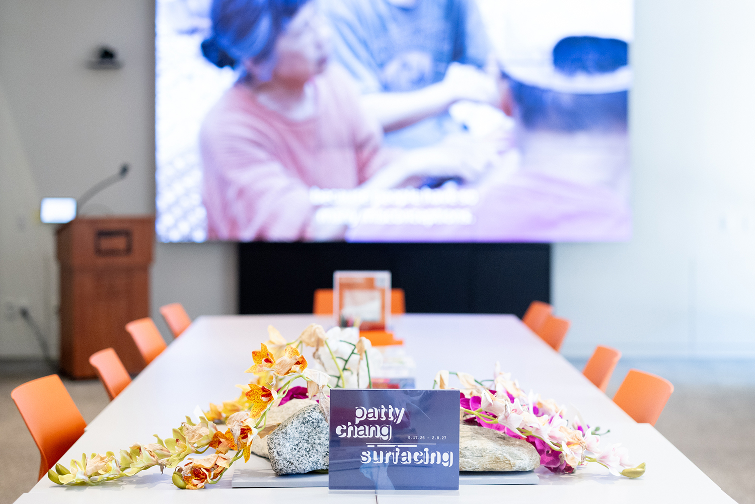 A conference table holds orchids, stones, and a small exhibition card, with a blurred video screen behind it.