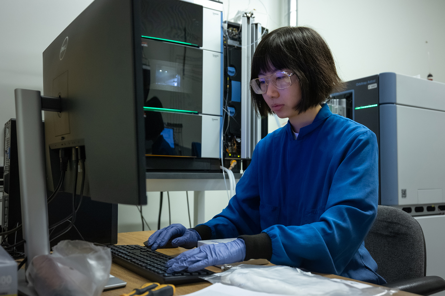 Nina Zhao types at a computer in front of large laboratory analysis instruments.