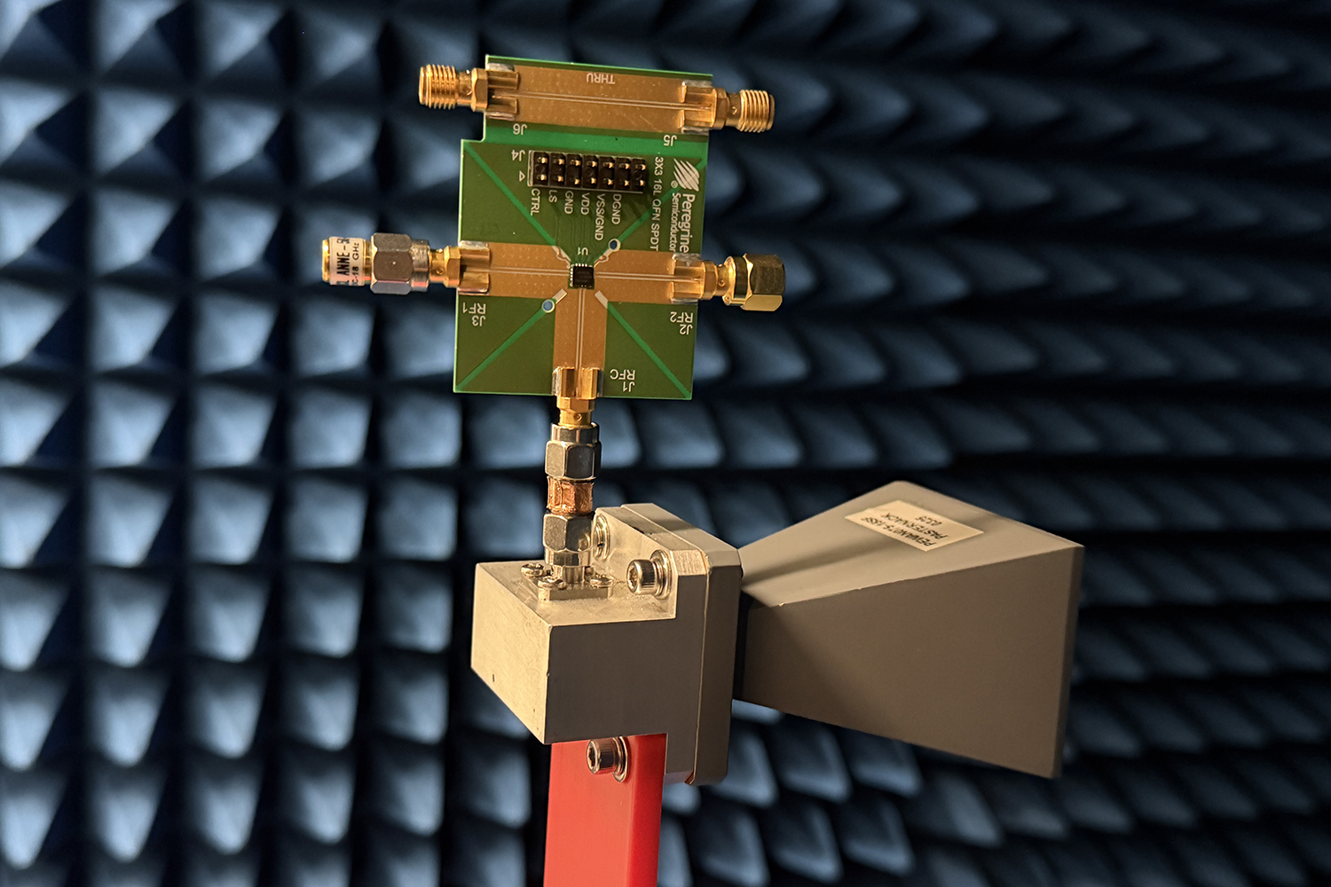 Green circuit board with gold connectors mounted on a horn antenna inside a chamber lined with blue foam cones.