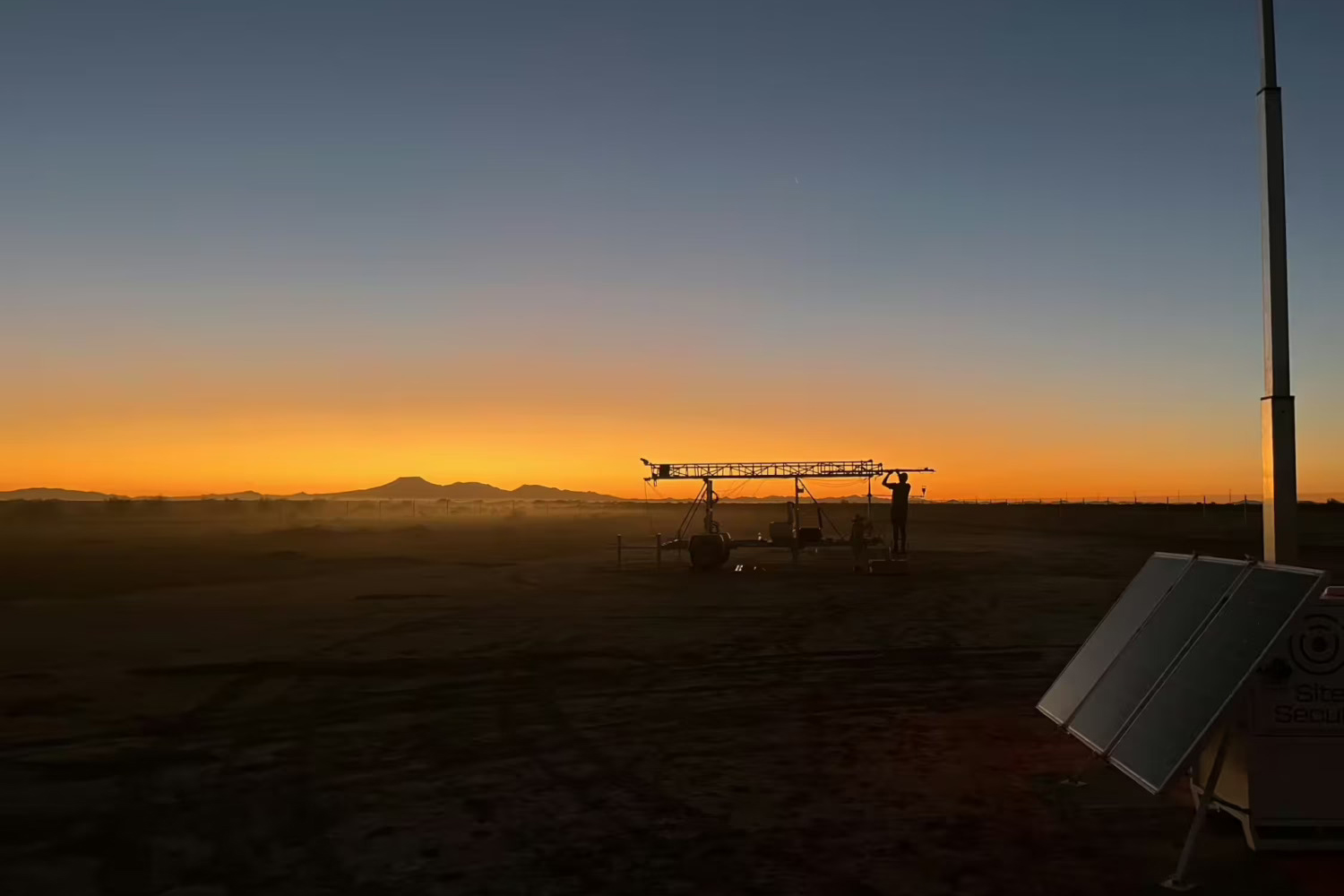 Taylor Adams, silhouetted by the dim light of the sun, works on a mobile equipment trailer in a desert field, with solar panels in the foreground and mountains beyond.