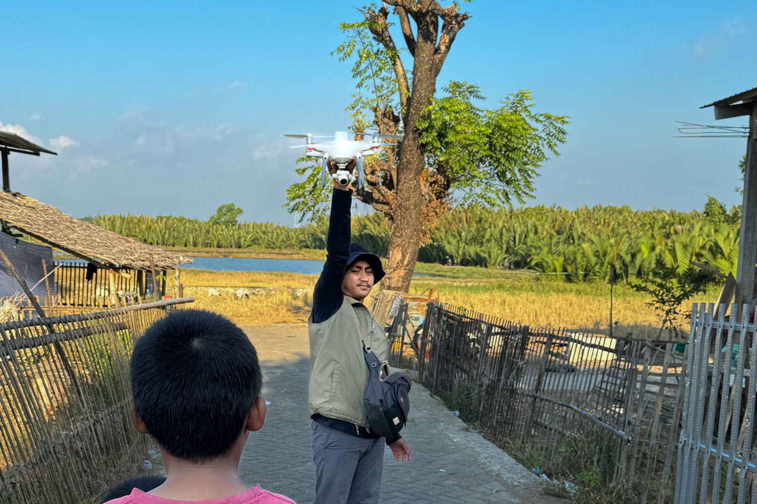 Man holds a drone above his head while a child watches on a rural path near fields and water.