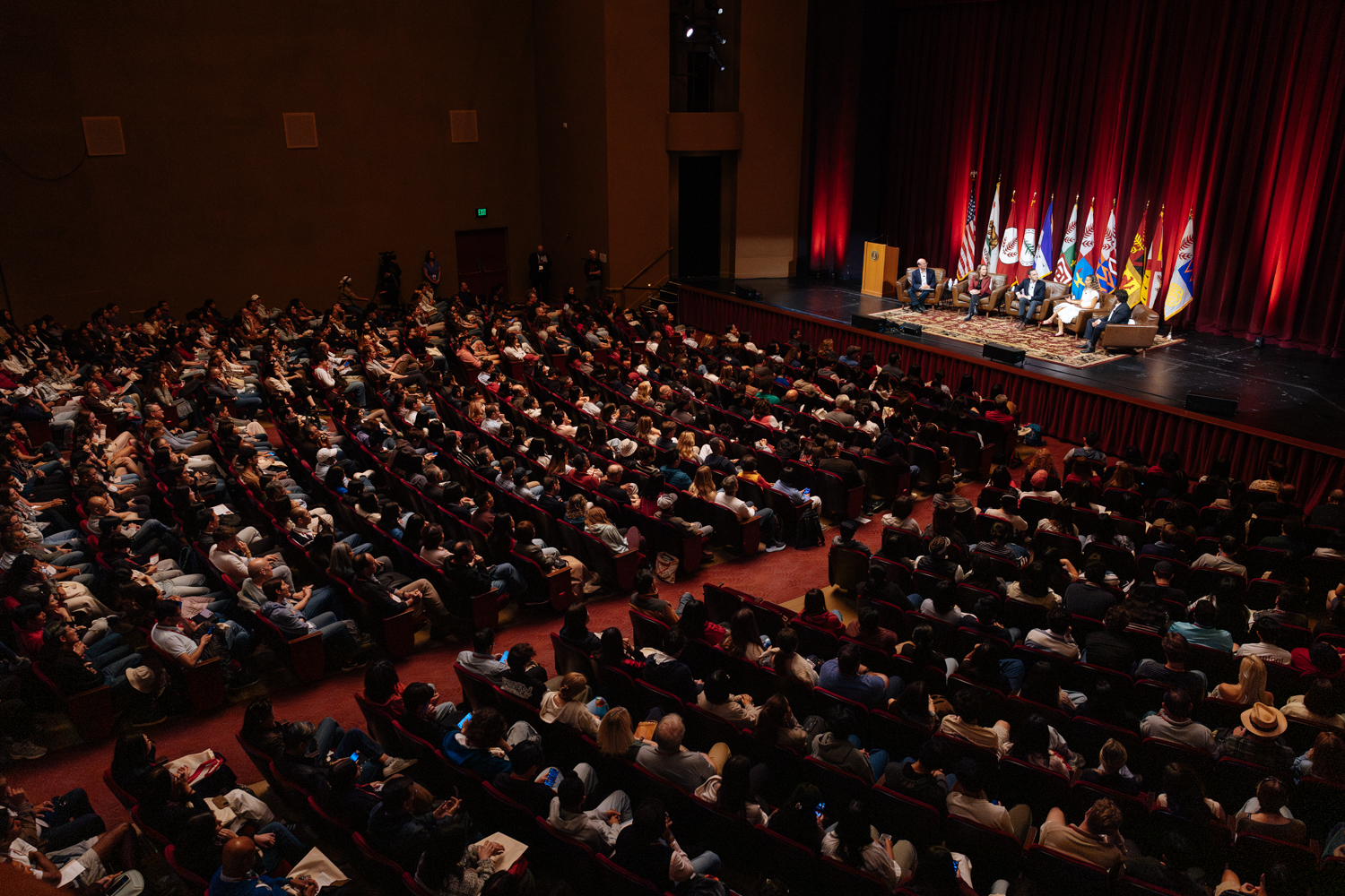 A large audience attending a panel discussion on stage, with five speakers seated and flags displayed in the background.