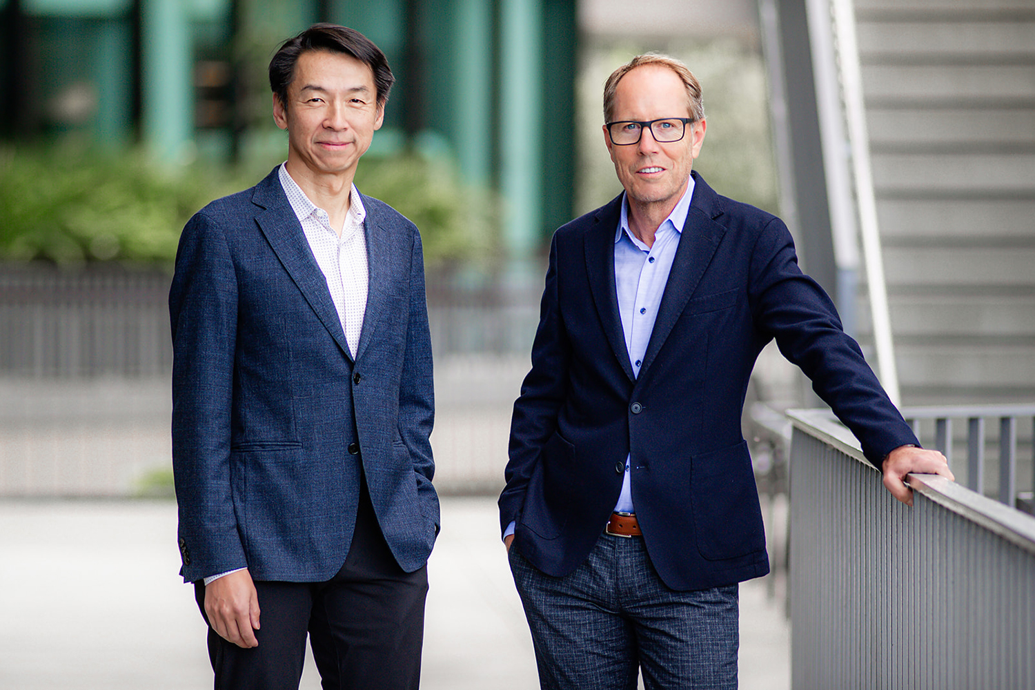 Kang Shen and Tony Wyss-Coray in business attire stand side by side outdoors, with a building and greenery in the background.