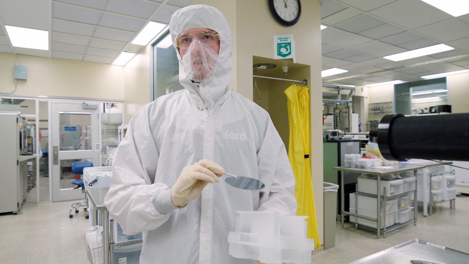 A lab worker in protective gear holds an item with prongs and a container while standing in a cleanroom environment.