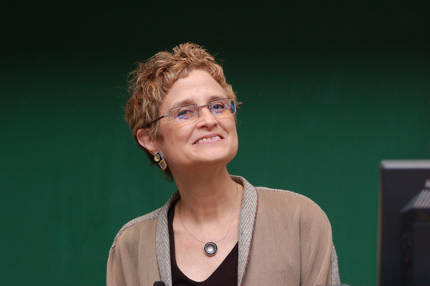Michele Barry wearing a beige jacket and round pendant looking up against a green background with a monitor to the right.