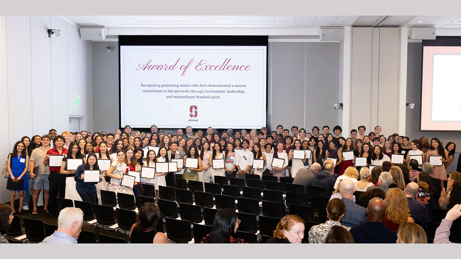 A large group of students stands together in a hall, holding certificates at an award ceremony celebrating their achievements.