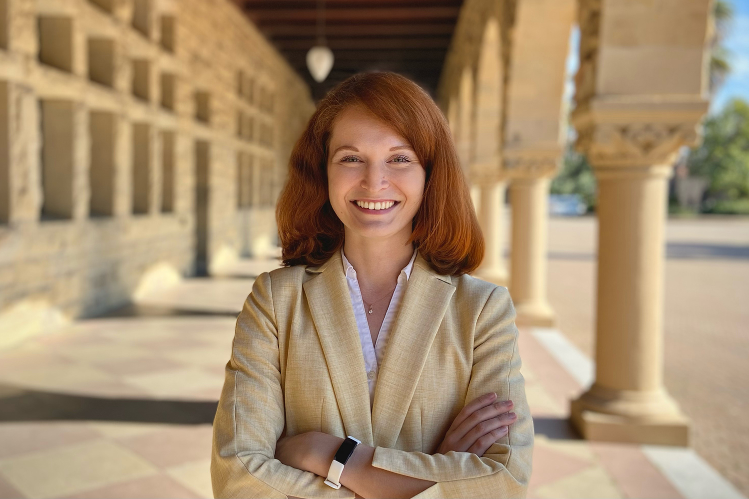 Petrova with red hair in beige blazer standing with arms crossed in an arcade of the Main Quad.