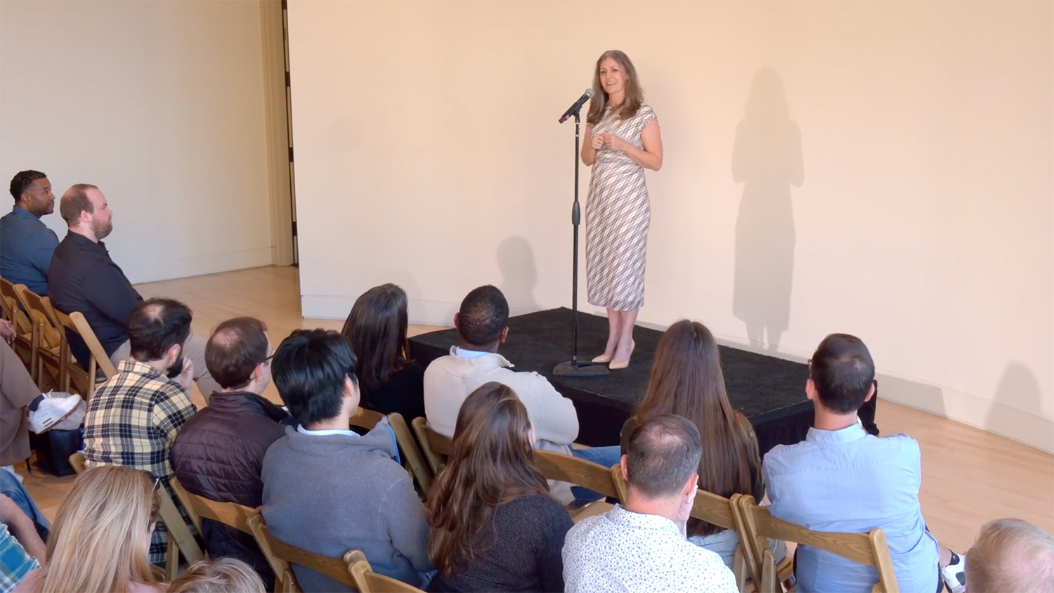 A woman in a patterned dress stands on stage holding a microphone, addressing an audience seated on wooden chairs.