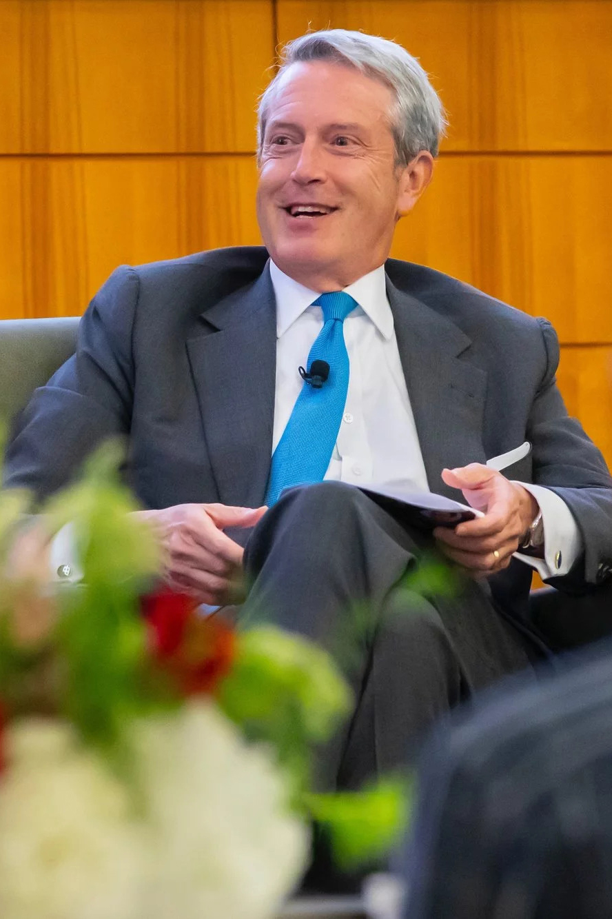 Randal Quarles in a suit and blue tie sits in a chair, holding notes and engaged in discussion.