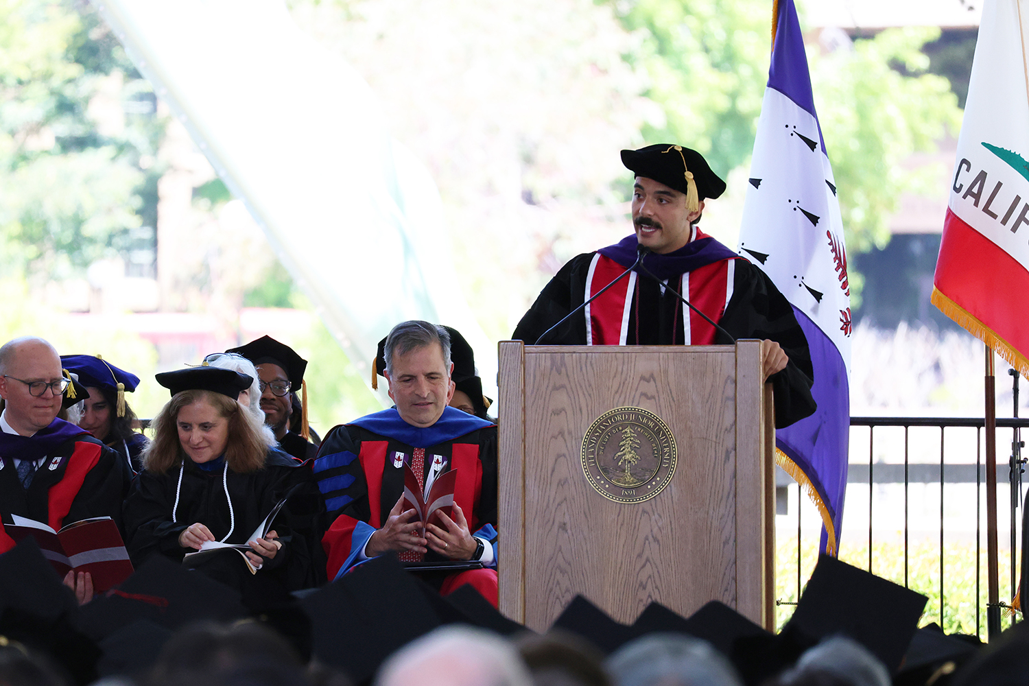 Graduate Jared James Hrebenar addresses the audience from a podium at a graduation ceremony, with faculty and flags in the background.